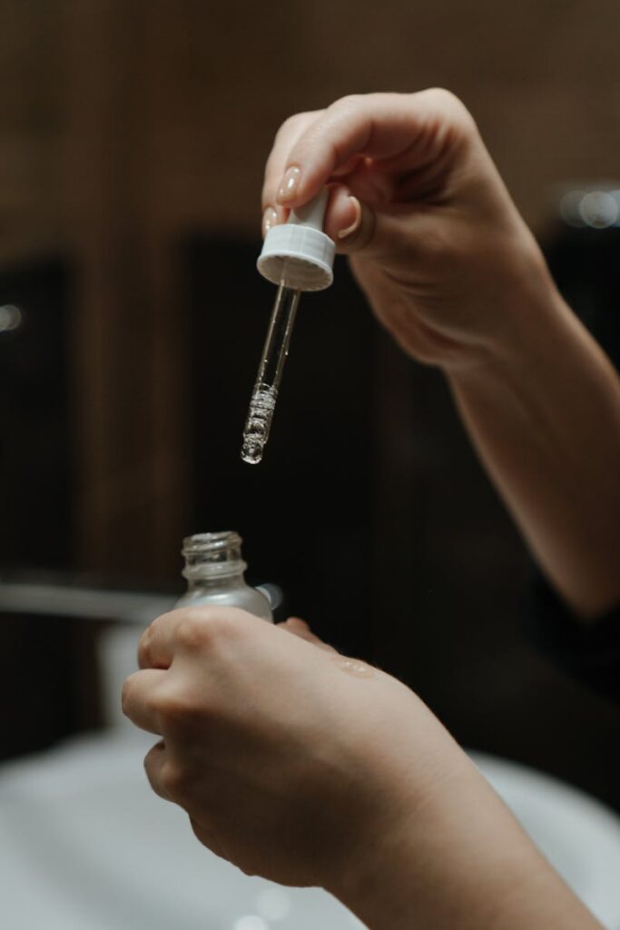 Hands holding a serum dropper applying skincare product in a bathroom setting.