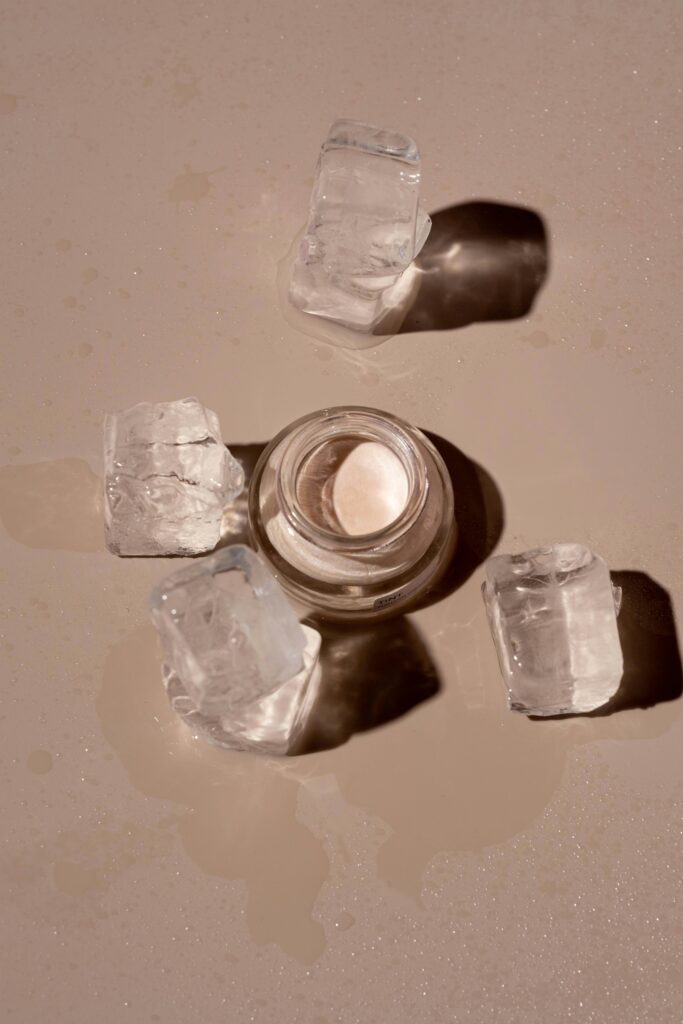 Flat lay of a skincare product surrounded by ice cubes on a textured surface.